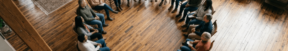 Diverse group of people sitting on wooden chairs in a circle in a room with wooden floors